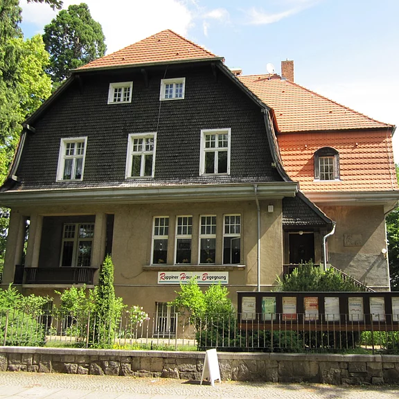 Zweistöckiges, historisches Haus mit dunkler Schindelfassade oben und rotem Ziegeldach. Über einer Eingangsterrasse, vor kleinem Garten und Zaun, hängt ein Banner mit der Aufschrift „Haus der Begegnung“.