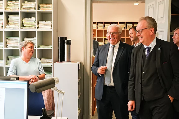 Eine Frau und mehrere Männer stehen in einem Büro. Die Frau lächelt, während die Männer, einer in Anzug und Krawatte, fröhlich lachen und miteinander sprechen. Im Hintergrund sind Aktenordner sichtbar, die auf Regalen stehen. Ein Computer ist ebenfalls im Bild zu sehen.