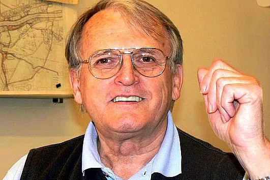 Porträt eines älteren Mannes mit Brille, der freundlich in die Kamera lächelt und die rechte Hand in einer betonten Geste hebt. Neutrale Innenraumszene im Hintergrund.