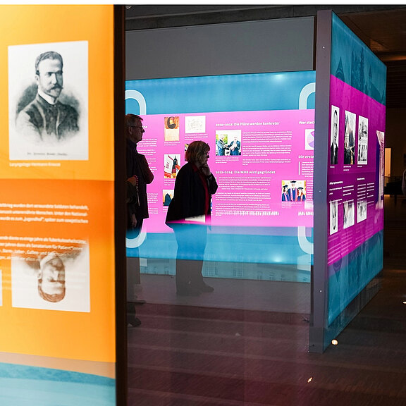 Die Abbildung zeigt eine Ausstellung mit farbigen Informationswänden und Portraits. Besucher stehen vor den Wänden, die interessante Inhalte präsentieren. Der Fokus liegt auf der interaktiven Gestaltung und den dargestellten historischen Informationen.