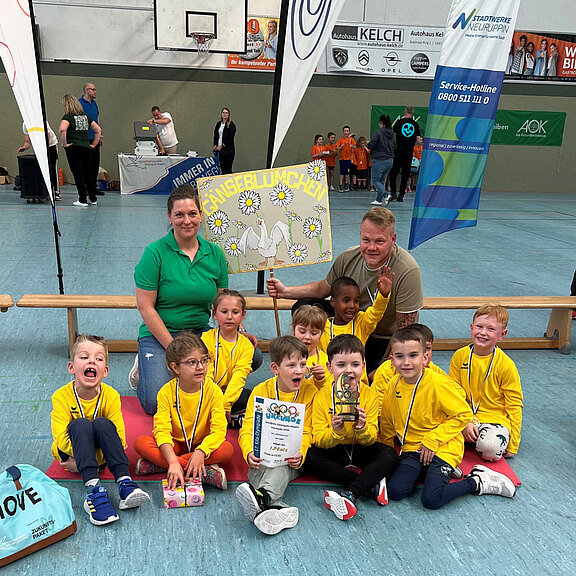 Eine Gruppe von Kindern in gelben T-Shirts sitzt auf dem Boden und hält eine Trophäe sowie ein Schild mit der Aufschrift "Wünsche blühen". Zwei Erwachsene, eine Frau und ein Mann, sitzen hinter den Kindern und lächeln. Die Atmosphäre ist festlich und erfreulich.