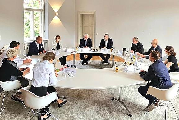 Eine Gruppe von Menschen sitzt um einen großen, ovalen Tisch in einem hellen Raum. Sie scheinen in eine Diskussion vertieft zu sein. Einige Getränke und Snacks sind auf dem Tisch verteilt. Der Fokus liegt auf dem Austausch von Ideen und Meinungen.