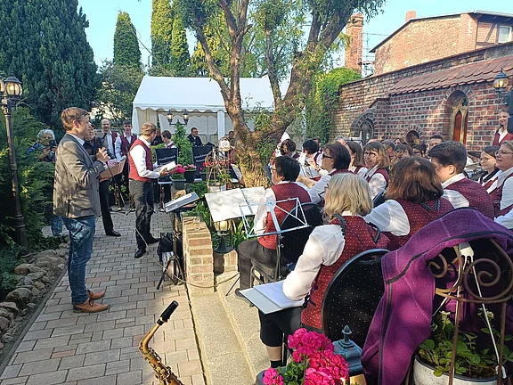 Ein Blasorchester spielt im Freien. Der Dirigent leitet die sitzenden Musikerinnen und Musiker an ihren Notenständern. Alle tragen einheitliche Westen. Der Auftritt findet in einem Gartenhof neben einer Backsteinmauer statt.