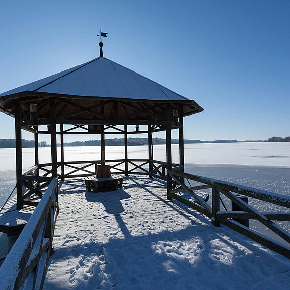 Ein hölzerner Pavillon steht auf einem gefrorenen See, umgeben von Schnee. Die Sonne scheint, und der klare Himmel verstärkt die kalte, ruhige Atmosphäre. Der Pavillon hat ein schräges Dach und eine offene Struktur mit Blick auf die umliegende Landschaft.