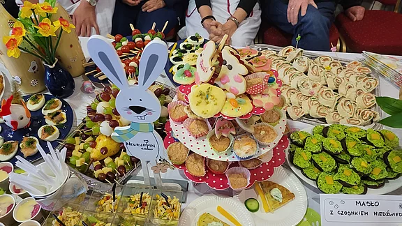 Eine festliche Essensauswahl auf einem Tisch, einschließlich bunter Leckereien wie Muffins, Obstspießen, Fingerfood und kleinen Desserts. In der Mitte steht ein dekorativer Hase, der die fröhliche Atmosphäre einer Feier unterstreicht.