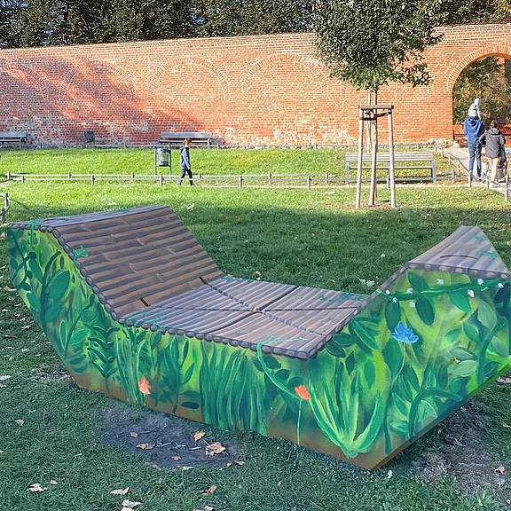 Wellenförmige Parkliege mit Holzlatten; Sockel bunt mit Gräsern und Blumen bemalt; steht auf Rasen in einem Park vor einer Ziegelmauer.