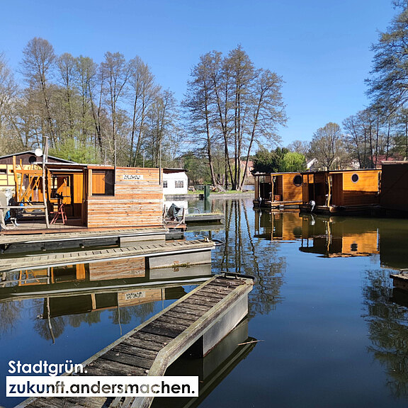 Eine friedliche Wasserlandschaft mit Holzbooten und einem Steg, umgeben von Bäumen. Die reflexive Oberfläche des Wassers spiegelt die Natur wider, während das Motto "Stadtgrün. zukunft.andersmachen." im Vordergrund sichtbar ist.