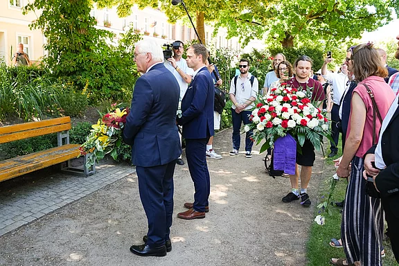 Zwei Männer in Anzügen stehen in einem Park und halten Blumensträuße. Eine Gruppe von Menschen ist im Hintergrund versammelt. Ein Mann trägt einen großen Blumenkorb. Es scheint eine Gedenkveranstaltung oder Zeremonie zu sein.