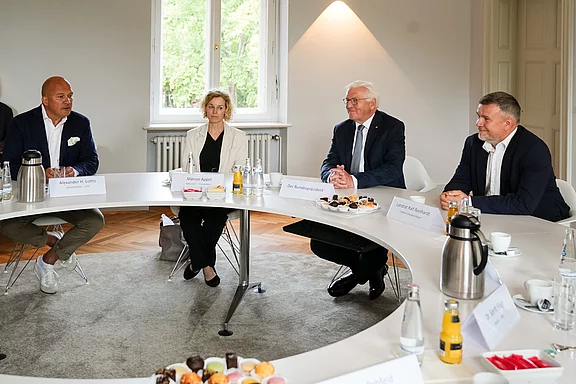 Ein runder Tisch mit mehreren Personen, die miteinander sprechen. In der Mitte befinden sich Getränke und Snacks. Der Raum ist hell erleuchtet und hat Fenster, die auf eine grüne Umgebung blicken. Die Atmosphäre wirkt formal und geschäftlich.