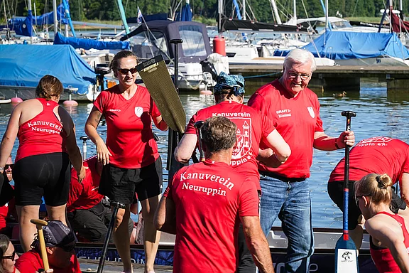 Eine Gruppe von Personen in roten Shirts mit der Aufschrift "Kanuverein Neuruppin" bereitet sich am Wasser auf das Paddeln vor. Einige tragen Paddel, andere steigen ins Boot. Im Hintergrund sind Boote im Hafen sichtbar.