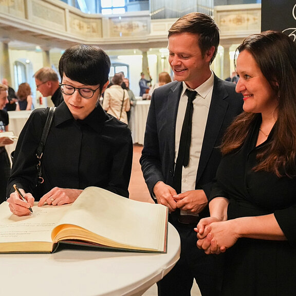 Eine Person mit Brille schreibt in ein großes Buch, während zwei weitere Personen zuschauen und lächeln. Die Szene findet in einem festlichen Raum mit anderen Anwesenden im Hintergrund statt.