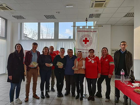 Gruppe von Freiwilligen steht in einem hellen Raum vor einem Banner der Croce Rossa Italiana. Mehrere halten Auszeichnungen in den Händen. Die Szene wirkt wie eine kleine Ehrung oder Dankesfeier.