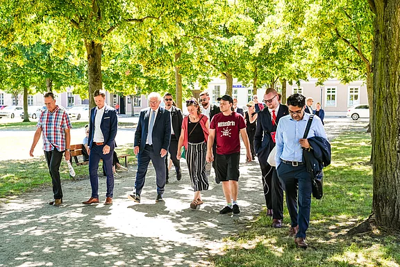 Eine Gruppe von sieben Personen geht auf einem schattigen Weg unter Bäumen. Sie tragen verschiedene Kleidung, von formellen Anzügen bis hin zu lässigen Outfits, und scheinen miteinander zu diskutieren. Die Szene vermittelt eine freundliche und gesellige Atmosphäre.