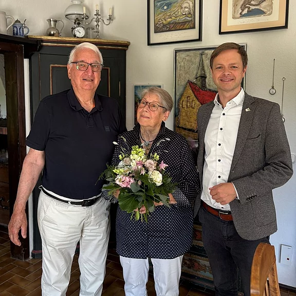 Drei Personen in einer Wohnung: In der Mitte hält eine ältere Frau einen Blumenstrauß und lächelt. Links und rechts stehen zwei Männer, einer leger, der andere im Sakko. Der Moment wirkt feierlich.