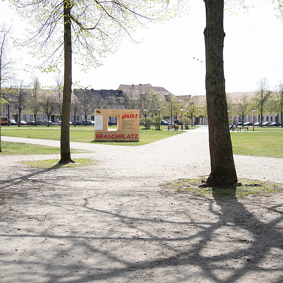 Ein sonniger Park mit einem großen grasbewachsenen Platz. In der Mitte steht ein auffälliges, orangefarbenes Schild mit der Aufschrift "Braschplatz". Umgeben von Bäumen und Gebäuden, lädt der Platz zum Verweilen ein.