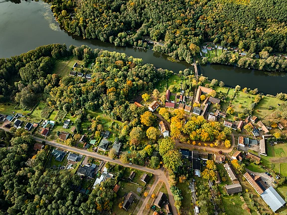 Luftaufnahme eines kleinen Dorfes umgeben von Wäldern und einem Fluss. Die Bäume zeigen herbstliche Farben, während die Häuser in einem grünen Landschaftsbild verteilt sind. Eine Straße verläuft durch das Dorf und verbindet die Gebäude.