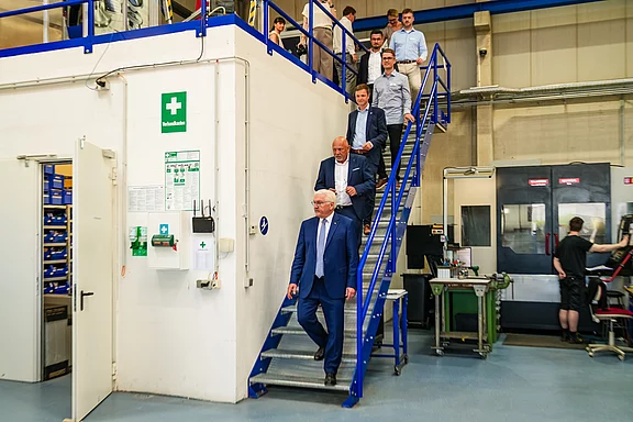 Eine Gruppe von fünf Männern, darunter ein Mann in einem blauen Anzug, geht eine Treppe hinunter in einem Produktionsgebäude. Im Hintergrund sind einige Arbeitsplätze und Maschinen zu sehen.