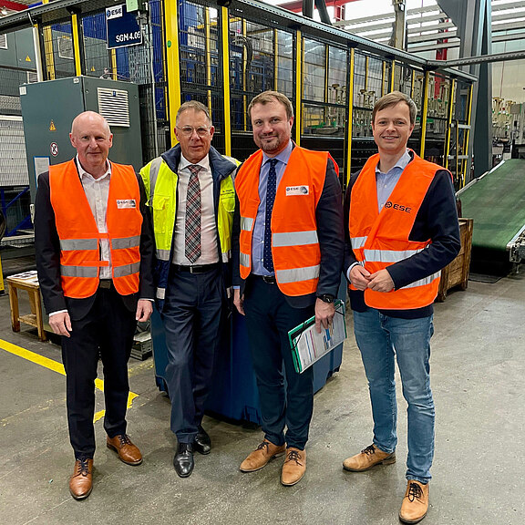 Vier Männer stehen in einer Industriehalle, tragen leuchtend orangefarbene Sicherheitswesten. Sie posieren gemeinsam für ein Foto. Im Hintergrund sind Maschinen und Ausrüstungen zu sehen, die auf eine industrielle Umgebung hinweisen.