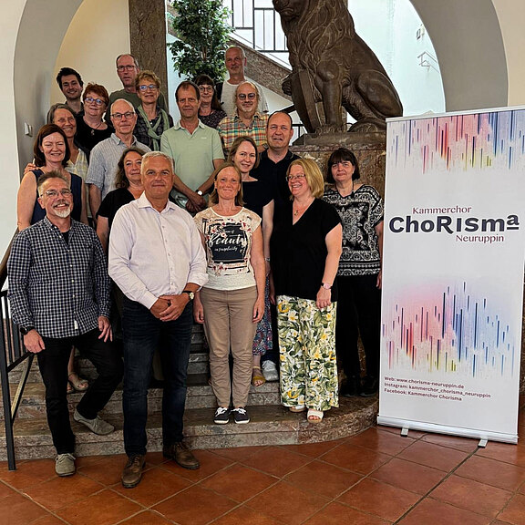 Eine Gruppe von Menschen steht auf einer Treppe vor einer Statue eines Löwen. Einige Personen tragen unterschiedliche Kleidungsstile. An der Seite befindet sich ein Plakat des Kammerchors ChoRiSma Neuruppin. Die Atmosphäre wirkt freundlich und einladend.