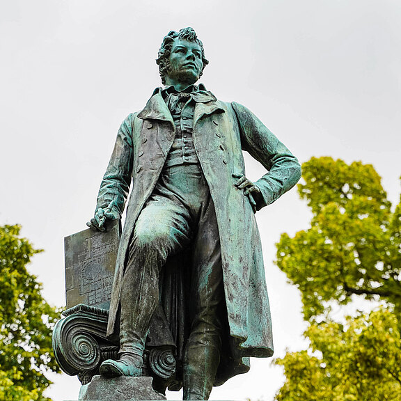 Eine Statue eines Mannes in historischer Kleidung steht dominant in der Mitte. Er hat eine Hand in die Hüfte gestützt und blickt nach vorne. Im Hintergrund sind grüne Bäume sichtbar. Die Statue vermittelt eine würdige und nachdenkliche Stimmung.