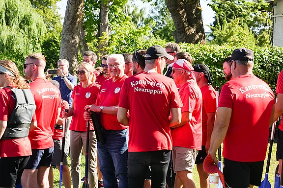 Eine Gruppe von Menschen in roten T-Shirts mit der Aufschrift "Kantorei Neuruppin" versammelt sich im Freien. Sie lächeln und scheinen auf eine Veranstaltung oder ein Team-Ereignis vorbereitet zu sein. Bäume und ein geschäftiger Hintergrund sind zu sehen.