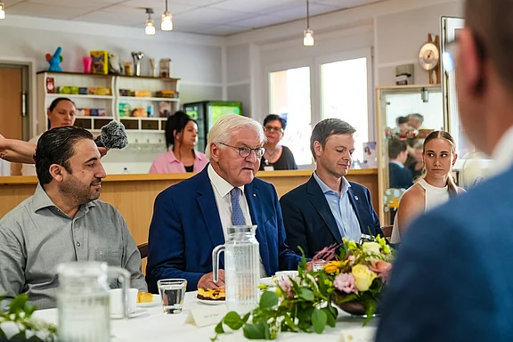 Eine Gruppe von Personen sitzt an einem gedeckten Tisch in einem hellen Raum. Im Mittelpunkt steht ein älterer Mann mit Brille, der einen blauen Anzug trägt. Es gibt Blumen und Getränke auf dem Tisch, und einige Personen im Hintergrund unterhalten sich.