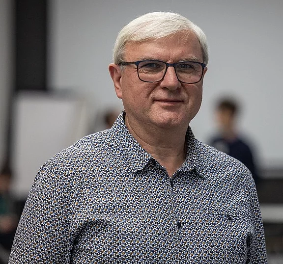 Ein Mann mit kurzen, grauen Haaren und einer Sehhilfe trägt ein gemustertes Hemd. Er steht im Vordergrund, während unscharfe Personen im Hintergrund zu sehen sind. Die Umgebung wirkt wie ein Veranstaltungsraum.