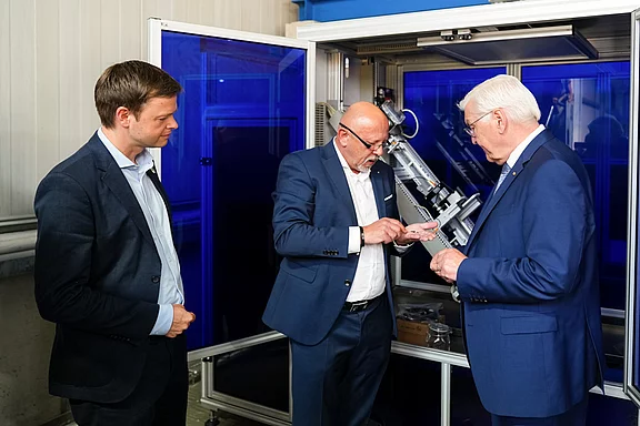 Drei Männer stehen in einem modernen industriellen Umfeld. Einer von ihnen zeigt etwas auf einem Display, während die anderen beiden aufmerksam zuschauen. Sie tragen formelle Kleidung und wirken in ein Gespräch vertieft. Der Hintergrund ist blau erleuchtet.
