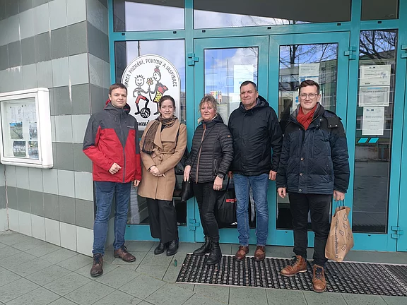 Fünf Erwachsene stehen lächelnd vor einem Gebäudeeingang mit türkisfarbenen Türen. Neben ihnen hängt ein rundes Logo mit Comic-Kindern, das auf eine Bildungseinrichtung hindeutet. Gruppenfoto bei kühlem Wetter, alle tragen Jacken.