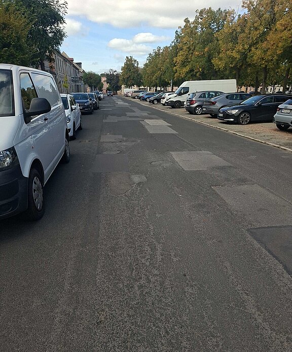Breite städtische Straße mit geparkten Autos beidseitig; Fahrbahn zeigt zahlreiche ausgebesserte Stellen und Flickenteppich aus Asphalt; kaum Verkehr; rechts Baumreihe, links Häuserfront; helles Tageslicht, leichter Wolkenhimmel.