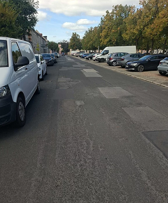 Breite städtische Straße mit geparkten Autos beidseitig; Fahrbahn zeigt zahlreiche ausgebesserte Stellen und Flickenteppich aus Asphalt; kaum Verkehr; rechts Baumreihe, links Häuserfront; helles Tageslicht, leichter Wolkenhimmel.