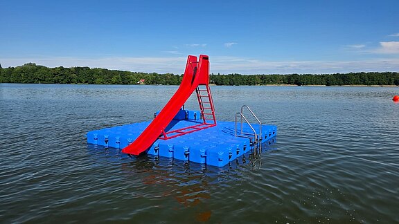 Blaue schwimmende Plattform mit roter Rutsche und Leiter auf einem See; dahinter bewaldetes Ufer und klarer Himmel.