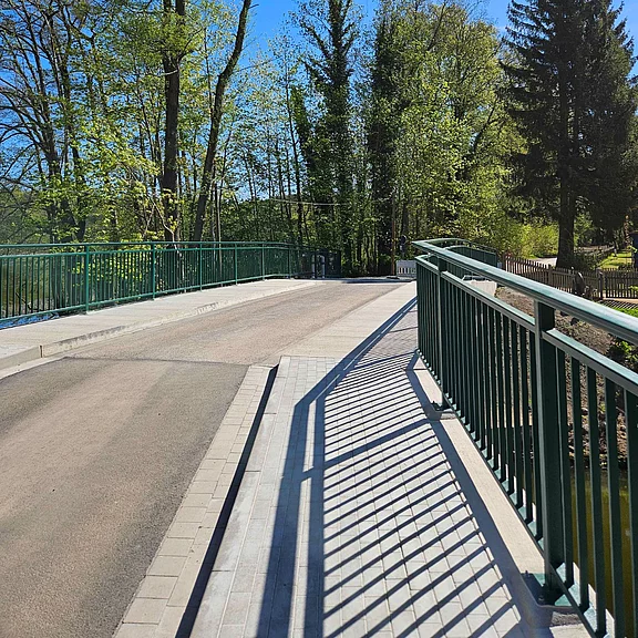 Sonnige Brücke mit grünem Geländer; rechts ein Fußweg, links die Fahrbahn. Lange Schatten der Geländer liegen auf dem Weg. Umgeben von frühlingsgrünen Bäumen unter blauem Himmel.
