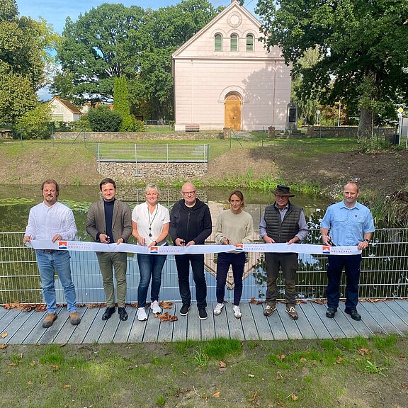Acht Personen stehen nebeneinander auf einer Holzplattform und schneiden gemeinsam ein langes Band durch – eine Einweihungsfeier. Hinter ihnen ein Geländer an einem kleinen Gewässer, dahinter Grünflächen und ein helles historisch wirkendes Gebäude.