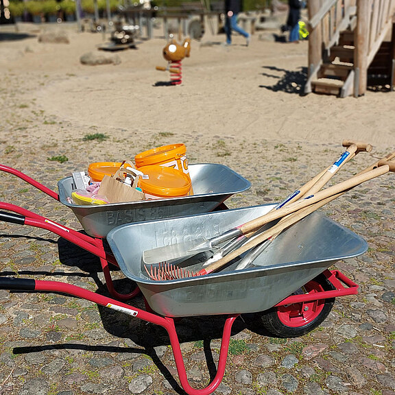Zwei Schubkarren stehen auf einem Spielplatz. Sie sind mit verschiedenen Werkzeugen ausgestattet, darunter Schaufeln und eine Harke. In einem der Schubkarren befinden sich orangefarbene Farbdosen. Der Hintergrund zeigt eine Spielgerätezone mit weiteren Besuchern.