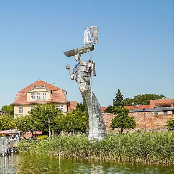 Große, silbern glänzende Skulptur einer Person, die einen Gleiter über dem Kopf hält. Die Figur steht am Ufer zwischen Schilf; im Hintergrund Gebäude, klarer blauer Himmel.