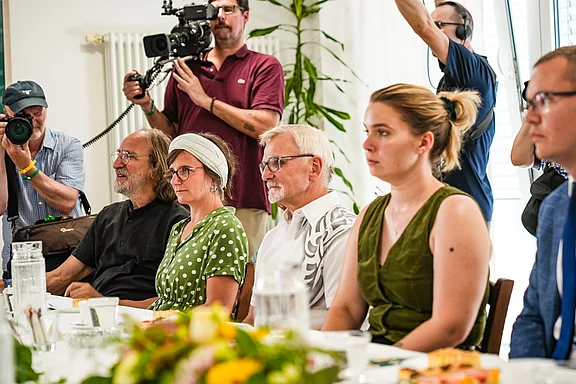 Eine Gruppe von fünf Personen sitzt an einem Tisch und hört aufmerksam zu. Sie sind verschieden gekleidet; eine Person trägt ein grünes Kleid mit weißen Punkten, eine andere ein dunkelgrünes Outfit. Im Hintergrund sind Kameraleute sichtbar, die die Szene filmen.