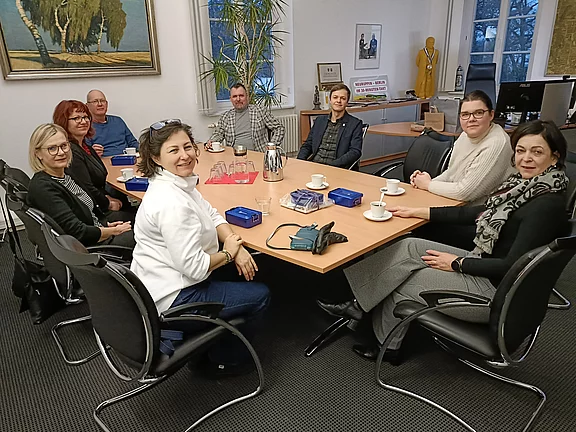 Eine Gruppe von acht Personen sitzt um einen Tisch in einem Meetingraum. Einige Tassen und Kaffeekannen sind auf dem Tisch, und im Hintergrund sind Kunstwerke sichtbar. Die Teilnehmer wirken in ein Gespräch vertieft und lächeln.