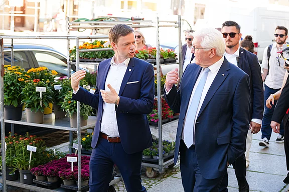 Zwei Männer, einer in Anzug und der andere in schicker Kleidung, unterhalten sich auf einem Gehweg neben einem Blumenstand. Im Hintergrund sind weitere Personen zu sehen, die die Straße entlanggehen. Es ist ein sonniger Tag.