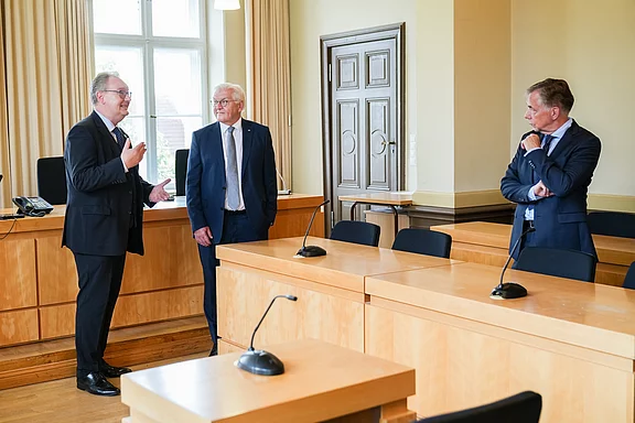 Drei Männer stehen in einem hellen Raum mit Holztischen. Einer spricht, während die anderen beiden aufmerksam zuhören. Der Raum wirkt formal und könnte ein Büro oder ein Besprechungsraum sein.