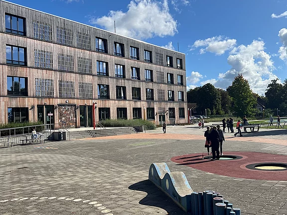 Bildbeschreibung: Eine moderne Schulgebäude mit Holzfassade steht auf einem weitläufigen Schulhof. Schüler und Schülerinnen bewegen sich auf dem Platz, der mit bunten Spielbereichen und Natur gestaltet ist. Ein klarer blauer Himmel mit wenigen Wolken ist im Hintergrund zu sehen.