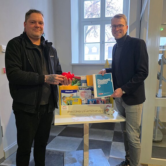 Zwei Männer stehen in einem Flur neben einem kleinen Tisch mit einer Spendenkiste voller Lebensmittel und Alltagsartikel. Der rechte hält eine Packung Studentenfutter hoch, der linke ein rotes Spielzeugauto. Sie lächeln in die Kamera.