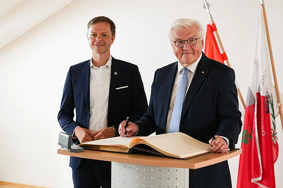 Der deutsche Bundespräsident steht an einem Tisch und signiert ein großes Buch. Neben ihm steht ein jüngerer Mann, der lächelt. Im Hintergrund sind Nationalflaggen sichtbar.