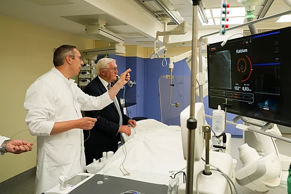 Ein Arzt in weißem Kittel erklärt einem älteren Mann etwas auf einem Bildschirm, der medizinische Daten anzeigt. Im Hintergrund ist eine Krankenhausumgebung zu sehen, mit medizinischer Ausstattung und einem Bett. Beide Personen scheinen interessiert an den Informationen auf dem Monitor zu sein.