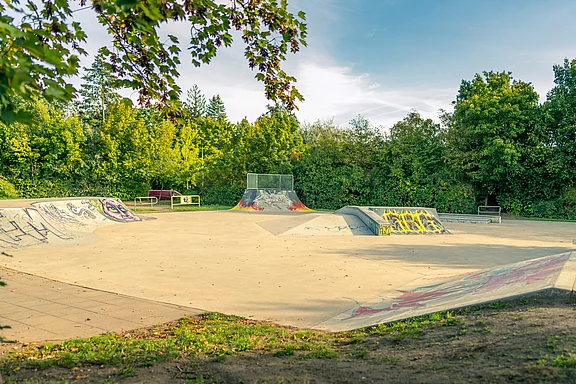 Leerer Skatepark im Freien mit mehreren Betonrampen und Quarterpipes, teils mit Graffiti, umgeben von dichtem Grün und Bäumen unter blauem Himmel.