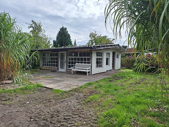 Kleines, einstöckiges Gartenhaus aus Holz mit großen Fenstern. Davor eine weiße Bank auf einer gepflasterten Terrasse. Umgeben von teils verwildertem Rasen und hohen Gräsern unter bedecktem Himmel.