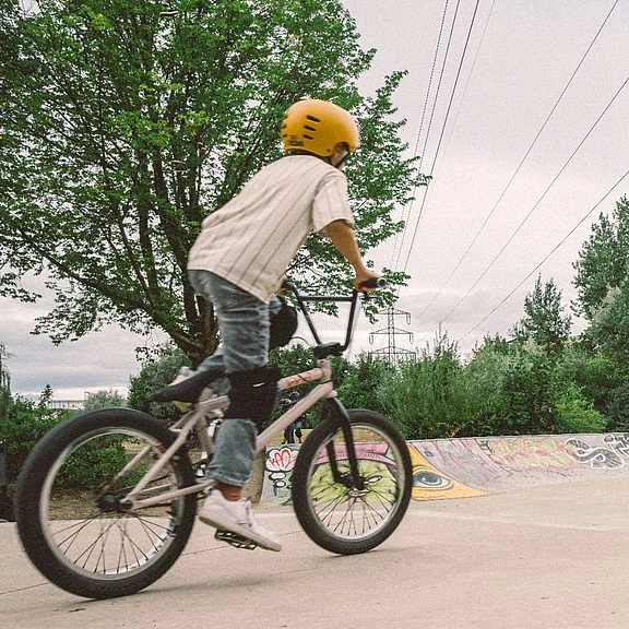 Kind mit gelbem Helm fährt auf einem BMX-Rad durch einen Skatepark, in Bewegung nach links. Knie- und Ellenbogenschoner, lässige Kleidung. Betonrampe und Bäume im Hintergrund, bewölkter Himmel. Szene vermittelt Outdoor-Aktivität und Dynamik.