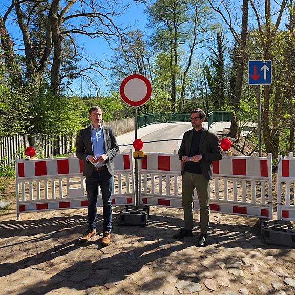 Zwei Männer stehen vor rot‑weißen Absperrgittern und einem „Durchfahrt verboten“-Schild an einer gesperrten Brücke. Dahinter eine schmale Straße im Grünen.