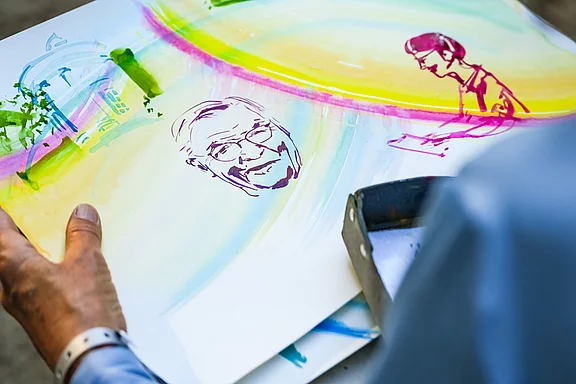 Eine Person hält ein künstlerisches Gemälde mit farbenfrohen Hintergründen. Auf dem Bild sind skizzenhafte Gesichter und Figuren zu sehen, die eine ruhige, kreative Stimmung vermitteln. Die Komposition vermittelt eine Verbindung zwischen Mensch und Kunst.