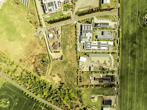 Senkrechte Luftaufnahme: rechts eine Reihe Gewerbegebäude mit Parkplätzen und Solardächern entlang einer Straße; links und unten weitläufige Felder und Grünflächen. Oben links ein Kreisverkehr. Deutlicher Übergang zwischen Industriegebiet und landwirtschaftlicher Fläche.